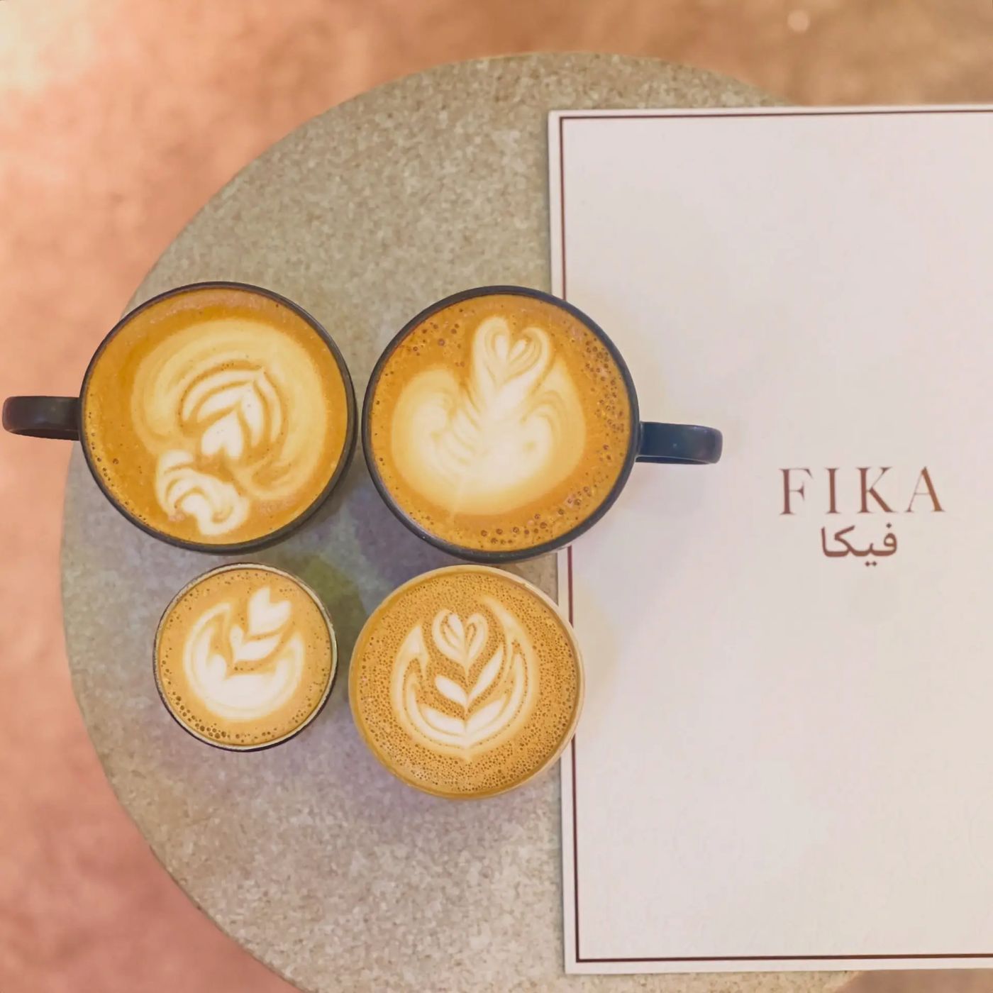 Four latte art coffees on a stone table beside a FIKA menu card